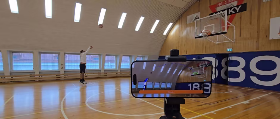 A person shooting a basketball in an indoor gymnasium, with a smartphone on a tripod in the foreground recording the action. The phone screen shows the live feed of the shot.